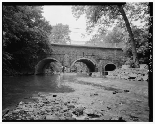 Black and white image of a stone bridge with two arches over a river, surrounded by trees and rocky riverbank.