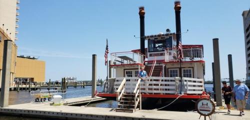A red and white riverboat docked at a marina, with people nearby and buildings in the background under a clear blue sky.