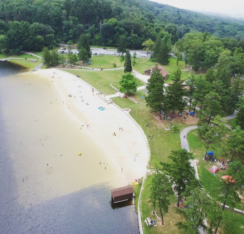 Aerial view of a sandy beach by a lake, surrounded by green trees and people enjoying the outdoors.
