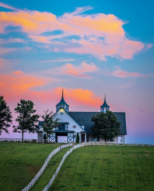 A picturesque barn with spires, set against a colorful sunset sky and surrounded by green fields and a white fence.