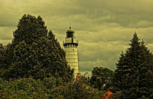 A tall lighthouse stands among lush green trees under a cloudy sky.
