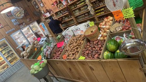 A rustic market display featuring various fresh vegetables, fruits, and jars, with colorful price tags and wooden crates.