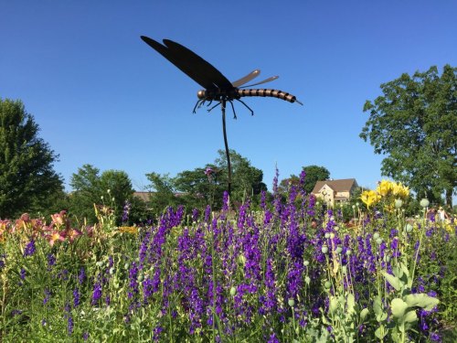 A large dragonfly sculpture perched on a stake amidst vibrant purple flowers under a clear blue sky.