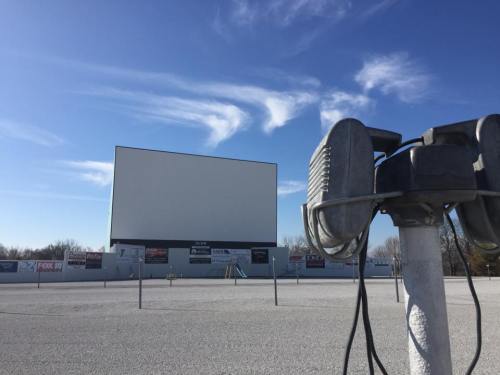 A blank outdoor movie screen stands in an empty lot under a blue sky with wispy clouds. A speaker is in the foreground.