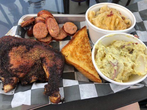A plate featuring smoked chicken, sausage, two sides of potato salad, and toasted bread on a checkered tray.