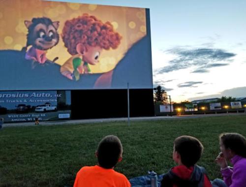 Children watching an animated movie on a large outdoor screen at dusk, with a colorful sky in the background.