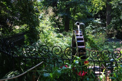 A vintage water wheel surrounded by lush greenery and a decorative iron fence in a serene garden setting.