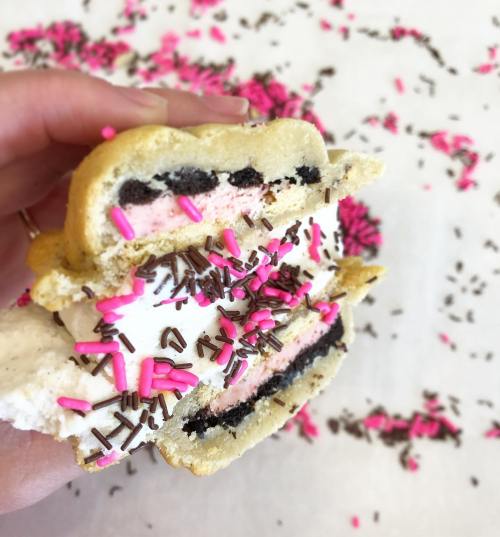 A hand holding a cookie sandwich filled with ice cream and sprinkles, surrounded by colorful sprinkles on a white surface.
