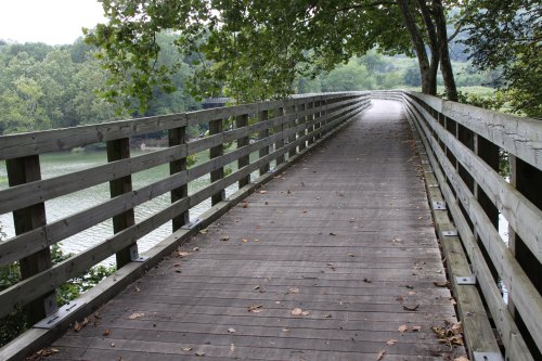 A wooden walkway lined with railings, leading through trees beside a calm river. Leaves scattered on the path.