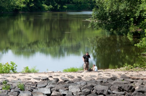 A man and a child stand by a calm river, surrounded by greenery and rocky shoreline on a sunny day.