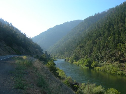 A serene river flows between lush green mountains under a clear blue sky, with a winding road nearby.