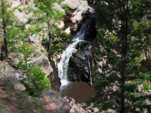A small waterfall cascades over rocks, surrounded by lush green trees in a natural setting.