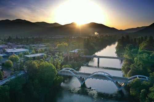 Aerial view of a river with two bridges, surrounded by trees and mountains, under a sunset sky.