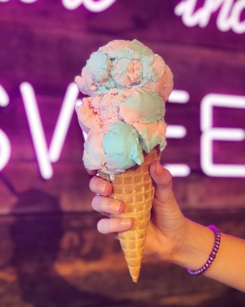 A hand holding a large ice cream cone with blue and pink scoops against a wooden background with neon lights.
