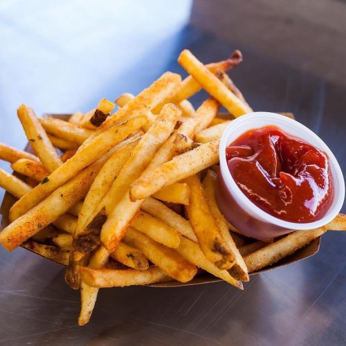 A serving of golden French fries in a paper tray, accompanied by a small cup of ketchup.