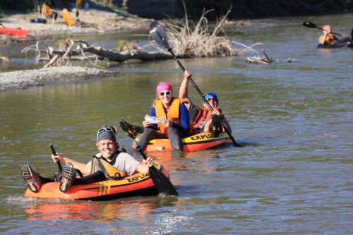 Three people kayaking on a river, smiling and enjoying the sunny day, with trees and other kayakers in the background.