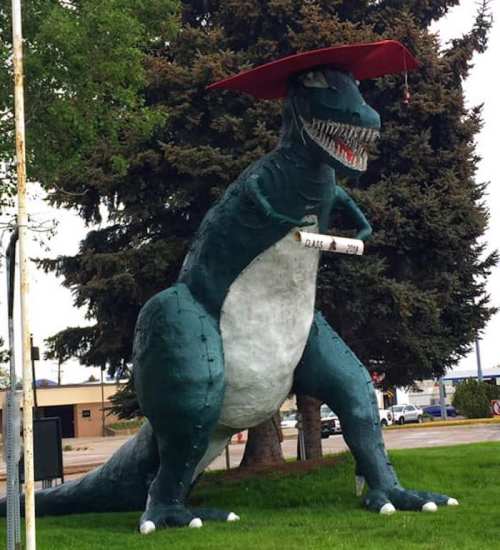 A large dinosaur statue wearing a graduation cap, holding a diploma, set against a backdrop of trees.