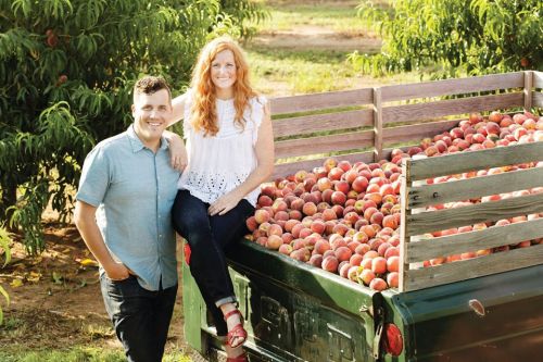 A man and woman pose beside a truck filled with fresh peaches in an orchard.