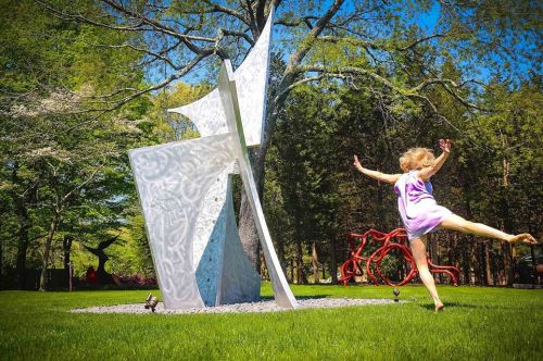 A dancer in a purple dress leaps joyfully near a large abstract sculpture in a green park.