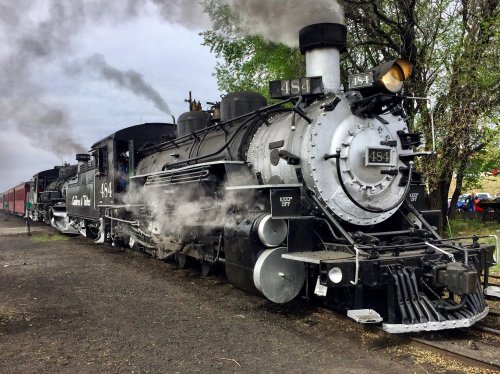 A vintage steam locomotive with billowing smoke, parked on a track surrounded by greenery.
