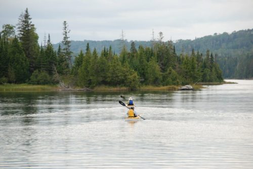 Two kayakers paddle on a calm lake surrounded by lush green trees and distant hills under a cloudy sky.