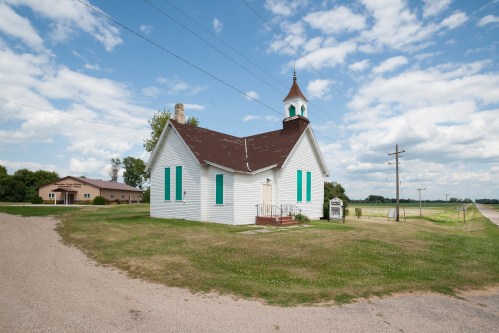 A small white church with green shutters, surrounded by grass and a dirt road, under a partly cloudy sky.