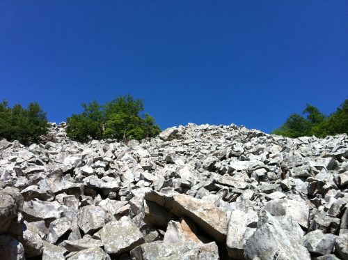A rocky hillside with scattered boulders and sparse greenery under a clear blue sky.