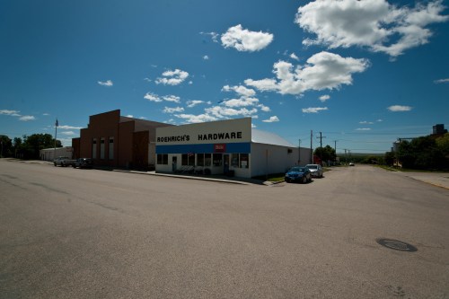A hardware store with a sign reading "Roerich's Hardware" on a sunny day, surrounded by a clear blue sky and parked cars.