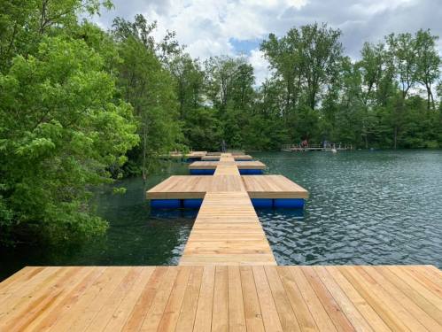 A wooden dock extends over a calm lake, surrounded by lush green trees under a cloudy sky.