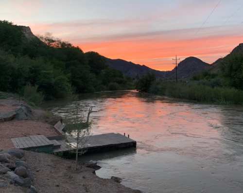A serene river at sunset, with vibrant colors in the sky and mountains in the background, framed by lush greenery.