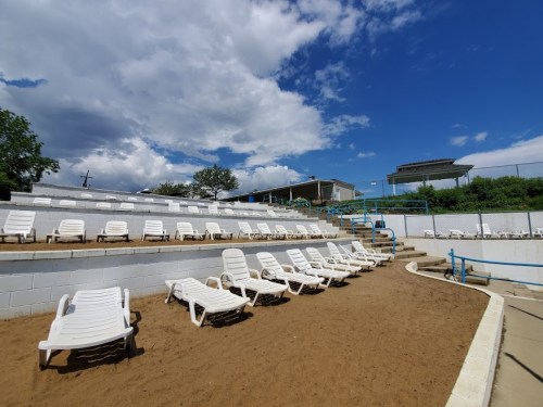 A sandy area with white lounge chairs arranged in rows under a blue sky with clouds.
