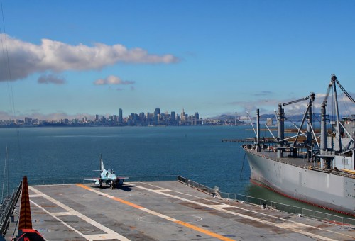 A view of a city skyline across the water, with a military aircraft on a ship's deck in the foreground.