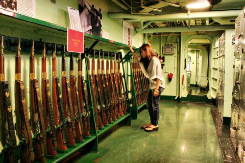 A woman examines a display of rifles in a military museum, surrounded by green walls and informational posters.