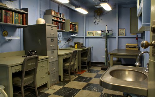 A small, dimly lit office with gray desks, filing cabinets, bookshelves, and a sink, featuring a checkered floor.