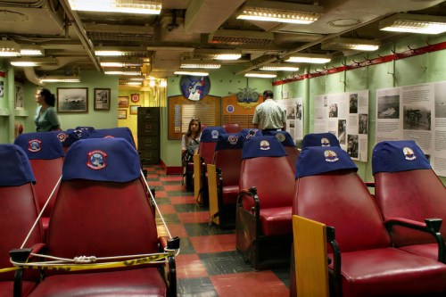Interior of a museum exhibit with red seats, informational displays on walls, and visitors exploring the space.