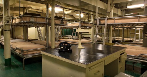 Interior of a dormitory with metal bunk beds, a desk with a vintage telephone, and green flooring.