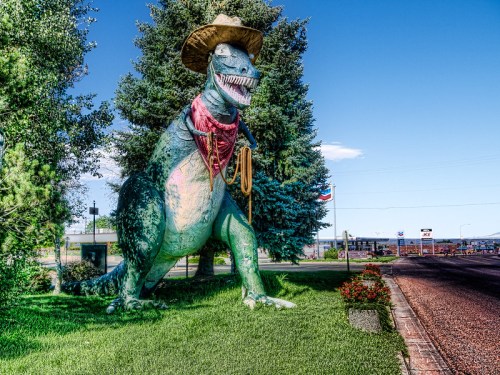 A large, colorful dinosaur statue wearing a cowboy hat and bandana, standing on grass near a road.