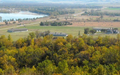 A scenic view of rolling hills, a river, and buildings surrounded by trees and fields with hay bales in the distance.