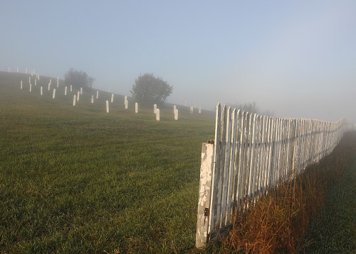 A foggy cemetery scene with white tombstones and a weathered wooden fence in a grassy landscape.