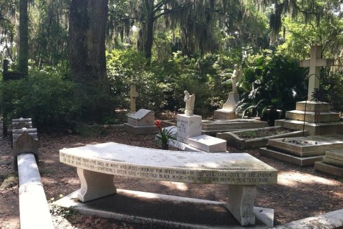 A serene cemetery scene featuring a stone bench surrounded by graves, trees, and lush greenery.