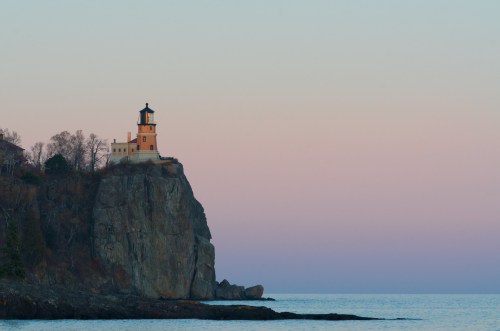 A lighthouse perched on a rocky cliff overlooking a calm sea at dusk, with a pastel sky in the background.