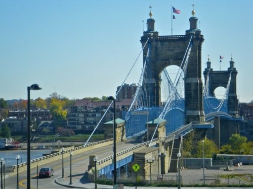 A blue suspension bridge with towers, flags, and a clear sky, spanning over a river with buildings in the background.