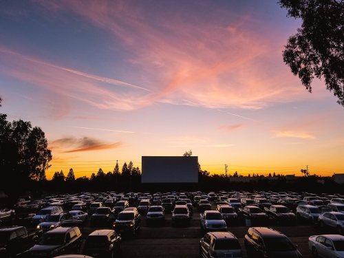 A drive-in movie theater at sunset, with cars parked in front of a large screen and colorful clouds in the sky.