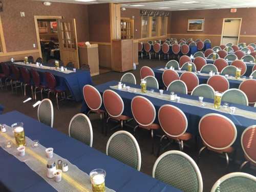 A spacious dining room with multiple tables set for a gathering, featuring round chairs and decorative table settings.