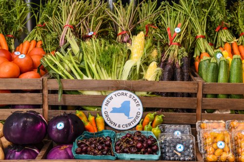 A wooden display of fresh vegetables, including carrots, tomatoes, corn, and eggplants, with a "New York State Grown" sign.