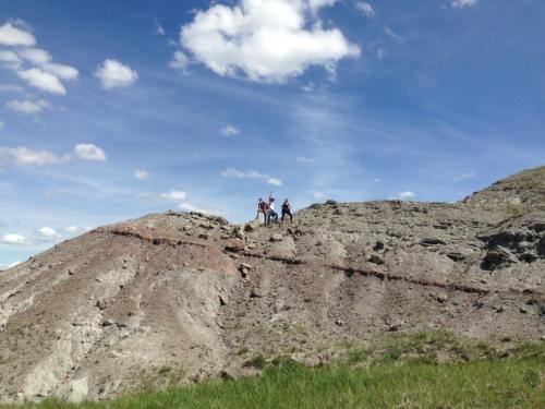 Two people stand on a rocky hillside under a blue sky with scattered clouds. Green grass is visible in the foreground.