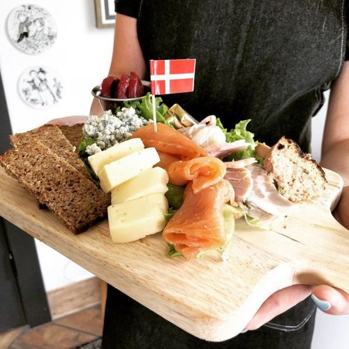 A wooden platter with assorted cheeses, smoked salmon, meats, and a small Danish flag, served on a bed of lettuce.