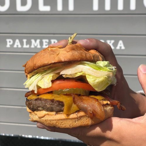 A hand holding a large burger with lettuce, tomato, pickles, cheese, bacon, and a toasted bun.