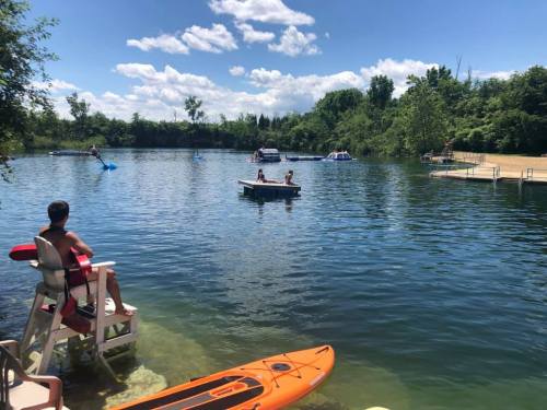 A sunny lake scene with people on a dock, a paddleboard, and a kayak, surrounded by trees and blue skies.