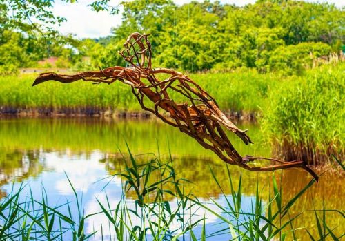A wooden sculpture of a figure in mid-air, surrounded by tall grass and a serene pond with lush greenery in the background.